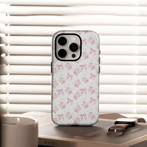 May include: A white phone case with a pattern of pink roses and bows. The case has a black border and a camera cutout. The phone case is on a table with a watch and a book.