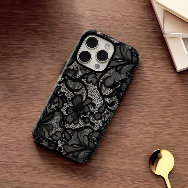 May include: Black and gray floral lace design phone case. The case features a detailed pattern of flowers and leaves. The phone case is on a wooden surface.