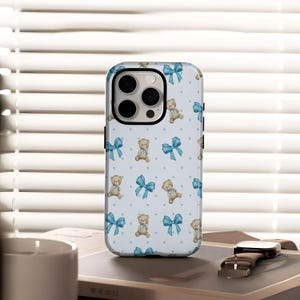 May include: A pale blue phone case adorned with a repeating pattern of teddy bears, blue bows, and small blue dots. The case has a black camera module. A watch and notebook are also visible.