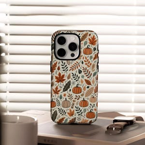 May include: A phone case with a fall-themed pattern. The case features a cream background with orange pumpkins, brown leaves, and green foliage. The case has a black border and a camera cutout.