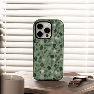 May include: A green phone case with a repeating pattern of pine cones, pine needles, and snowflakes. The case has a black border and a camera cutout. The phone case is sitting on a book next to a watch.