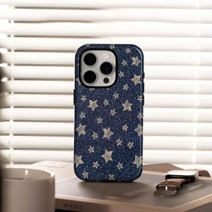 May include: A navy blue phone case with a pattern of small, glittery stars. The case has a black border and a camera cutout. A brown leather watch with a silver buckle rests on a book next to the phone.