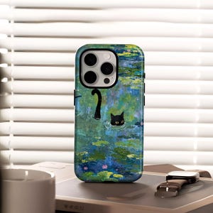 May include: A phone case with a design of a black cat swimming in a pond with lily pads. The case has an impressionistic style with blue, green, and pink hues. The phone case is on a table with a watch and a book.