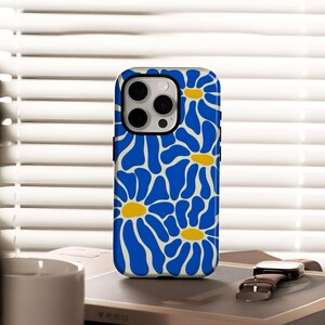 Könnte beinhalten: Eine Handyhülle mit blauem und weißem Blumenmuster. Das Case zeigt ein Muster aus blauen, gänseblümchenartigen Blumen mit gelben Zentren auf weißem Hintergrund. Die Handyhülle liegt auf einem Tisch mit einer Uhr und einem Buch.