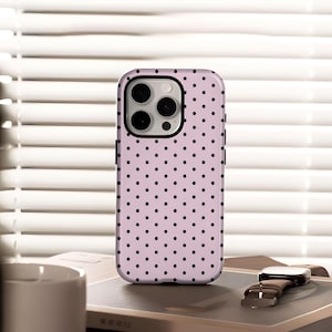 May include: A pale pink phone case featuring a black polka dot design. The case has a black edge and a camera aperture. A brown leather watch and a white mug are in the background.