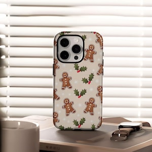 May include: A phone case with a festive gingerbread man pattern. The case features a repeating design of smiling gingerbread men, holly sprigs with red berries, and snowflakes on a beige background. The case is shown on a book with a watch.