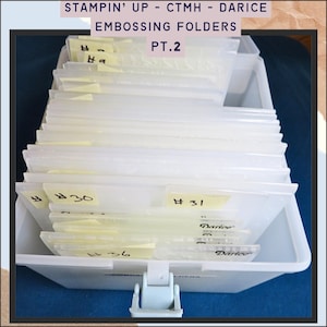 Può includere: Un contenitore di plastica bianco pieno di cartelle per goffratura trasparenti. Le cartelle sono etichettate con numeri scritti a mano su note adesive gialle. Il testo "STAMPIN' UP - CTMH - DARICE EMBOSSING FOLDERS PT.2" è in alto.