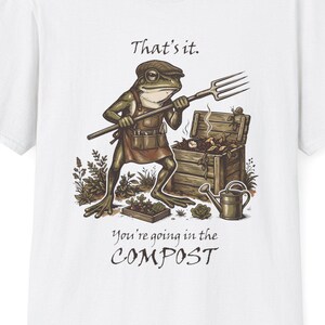 Op de afbeelding: Een wit t-shirt met een afbeelding van een groene kikker met een bruine pet en schort, die een riek vasthoudt naast een houten compostbak. De tekst luidt That's it. You're going in the COMPOST. Het ontwerp bevat ook een kleine gieter.