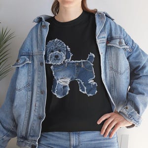May include: Black t-shirt featuring a graphic of a poodle made to look like it is wearing denim jeans. The poodle graphic is light blue and the jeans are distressed. The model is wearing a light blue denim jacket.