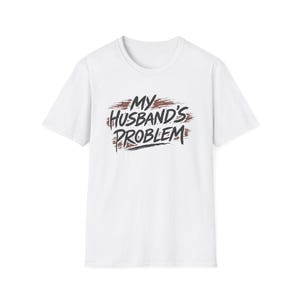 May include: White t-shirt with the phrase "MY HUSBAND'S PROBLEM" in a distressed, handwritten-style font. The text is in black and brown, with a brushstroke effect. The shirt has a crew neck and short sleeves.