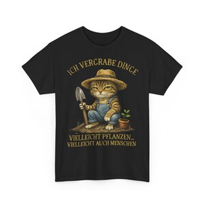 Könnte beinhalten: Schwarzes T-Shirt mit einer Cartoon-Katze, die einen Strohhut und eine Latzhose trägt und eine Schaufel hält. Der Text auf dem Shirt lautet "ICH VERGRABE DINGE" und "VIELLEICHT PFLANZEN... VIELLEICHT AUCH MENSCHEN."
