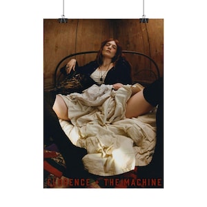 Op de afbeelding: Een poster met een vrouw die op een bed ligt, gekleed in een zwarte jas en een witte top. De poster heeft de tekst "FLORENCE + THE MACHINE" onderaan. De achtergrond is een houten muur.