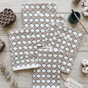 May include: Several hand-blocked cotton napkins with a scallop design in white, taupe, and green. The napkins are arranged on a wooden surface with a wooden block, paint, and cotton sprigs. A tag reads "HAND-BLOCKED COTTON."