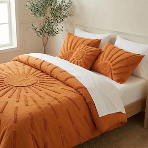 Op de afbeelding: Een oranje beddengoedset met een zonmotief. De set bevat een dekbedovertrek en drie sierkussens. Het zonmotief is gemaakt met verhoogde, getextureerde lijnen. Witte lakens contrasteren met het oranje dekbedovertrek.