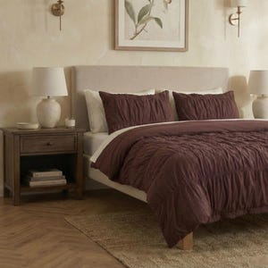 May include: A bedroom scene featuring a bed with a burgundy comforter and matching pillow shams. A wooden nightstand with a lamp and books sits beside the bed. The room has a neutral color palette with a framed botanical print.
