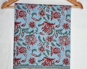 May include: A light blue fabric with a floral pattern of red and pink flowers and green leaves. The fabric is hanging on a wooden hanger. The bottom border features a white band with pink and blue bird and floral designs.