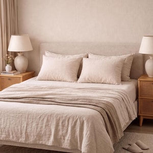May include: A bed with a beige linen duvet cover and two matching rectangular pillows. The bed features a light beige upholstered headboard. Two wooden nightstands with cane detailing sit on either side, each topped with a white lamp.