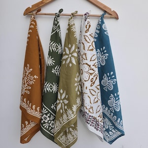 May include: Five colorful bandanas with white floral patterns hang from a wooden hanger. The bandanas are in shades of mustard yellow, olive green, white, and teal. Each bandana has a unique design, perfect for various uses.