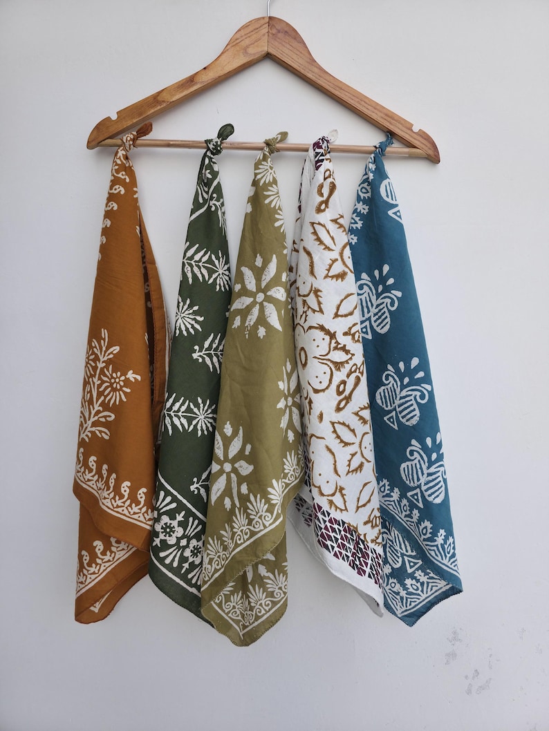 May include: Five colorful bandanas with white floral patterns hang from a wooden hanger. The bandanas are in shades of mustard yellow, olive green, white, and teal. Each bandana has a unique design, perfect for various uses.