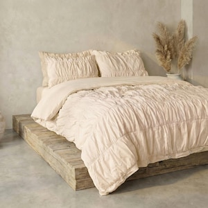 May include: A cream-colored bed set with a ruched duvet and pillow shams. The bedding is arranged on a rustic wooden platform bed frame. The room has a neutral color palette with a vase of dried plants.