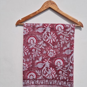 May include: A maroon and white floral patterned textile is displayed on a wooden hanger. The fabric features a repeating design of stylized flowers, leaves, and other botanical elements. The bottom edge has a striped border.