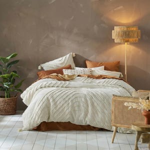 Könnte beinhalten: Ein Bett mit einer cremefarbenen Bettdecke und einem rostfarbenen Bettrock. Das Bett ist mit mehreren Kissen in Creme-, Weiß- und Rosttönen geschmückt. Eine gewebte Lampe und eine Topfpflanze befinden sich im Hintergrund und schaffen eine gemütliche Schlafzimmeratmosphäre.