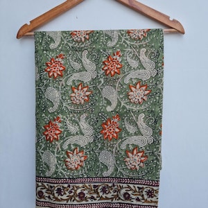 May include: A green fabric with a floral and paisley pattern, hanging from a wooden hanger. The design features orange flowers and off-white paisley shapes on a green background. The bottom edge has a cream border with a floral design.