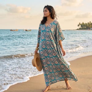 Puede incluir: Vestido largo turquesa con estampado floral, cuello en V y mangas cortas. El vestido se lleva en una playa de arena y se sostiene un sombrero de paja en la mano. El vestido presenta un patrón repetido de flores y hojas beige.