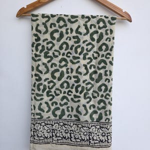 May include: A cream-colored scarf with a dark green leopard print pattern. The bottom edge features a black and white floral border. The scarf is hanging on a wooden hanger against a white background.