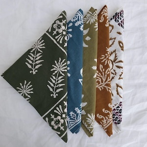May include: Five folded cloth napkins in a row, each with a different color and pattern. The colors include dark green, blue, olive green, brown, and white. Each napkin features a floral or geometric design in white or brown.