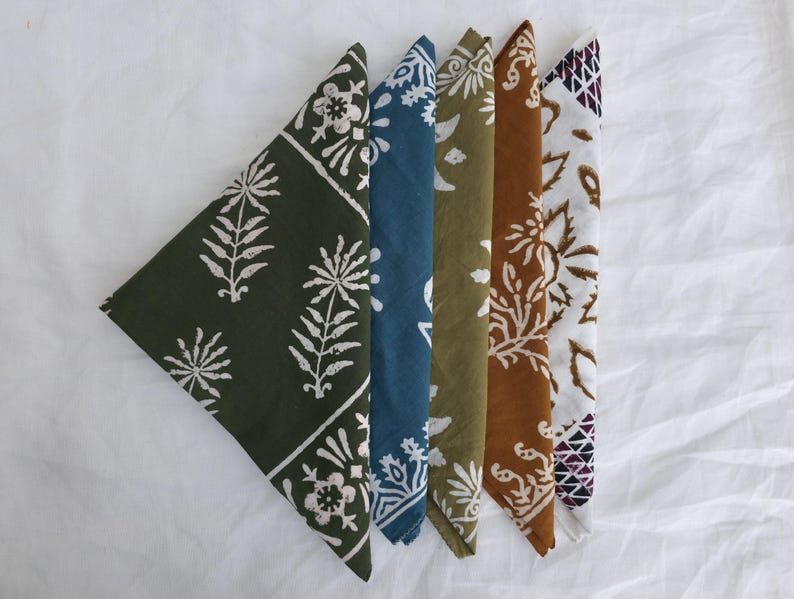 May include: Five folded cloth napkins in a row, each with a different color and pattern. The colors include dark green, blue, olive green, brown, and white. Each napkin features a floral or geometric design in white or brown.
