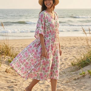 Può includere: Una donna indossa un abito bianco con stampa floreale rosa e un cappello di paglia su una spiaggia sabbiosa. L'abito ha uno scollo a V e una vita con coulisse. L'abito è al ginocchio e ha maniche corte. La donna indossa sandali marroni.