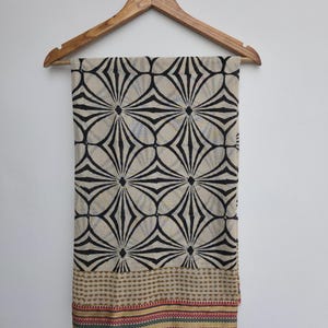 May include: A beige scarf with a black geometric pattern, hanging on a wooden hanger. The scarf features a repeating design of black lines radiating from central points. The bottom edge has a patterned border with brown, red, and green stripes.