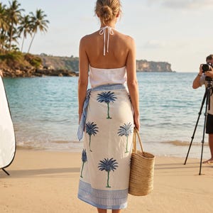 Może przedstawiać: Osoba stoi na piaszczystej plaży w białym topie typu halter i kremowym sarongu. Sarong ma niebieski nadruk z palmami i niebieską obw&oacute;dkę. Nosi plecioną torbę z wikliny. Tło obejmuje ocean, palmy i fotografa.