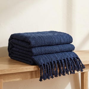May include: A folded navy blue throw blanket with a textured woven pattern and twisted fringe tassels along the edge. The fabric features a consistent deep blue color throughout, with a visible grid-like weave structure and knotted ends.