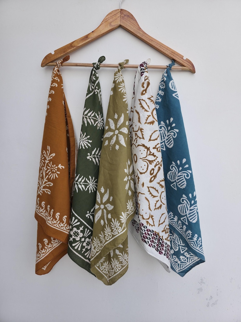May include: Five bandanas with white floral and bee designs hang from a wooden hanger. The bandanas are in shades of brown, green, olive, white, and teal. Each bandana has a different pattern, creating a colorful display.