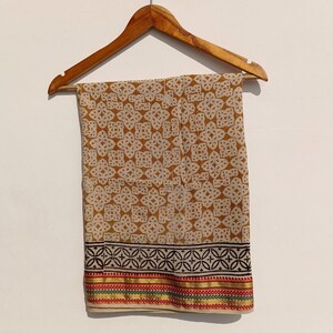 May include: A tan and white patterned fabric hanging on a wooden hanger. The fabric has a repeating floral design. The bottom border features a black geometric pattern with red, green, and yellow stripes.