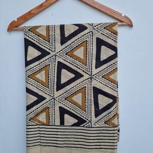 May include: A beige textile with a geometric pattern of black and brown triangles, hanging from a wooden hanger. The bottom edge features horizontal black stripes. The fabric is likely cotton or a similar material.