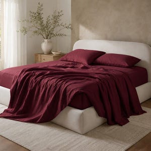 May include: A bed featuring a cream-coloured upholstered headboard and frame, dressed in deep burgundy bedding. The set includes a fitted sheet, a flat sheet draped over the side, and two matching rectangular pillows placed against the headboard.