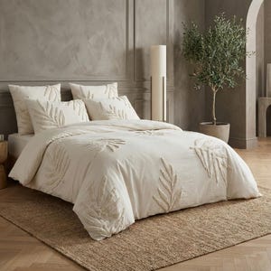 May include: Off-white bedding set with embroidered leaf patterns. The set includes a duvet cover and pillows, all with a textured design. The bed is on a natural fibre rug, creating a comfortable bedroom setting. The room has a neutral colour scheme.