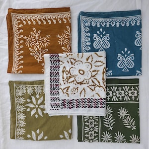 May include: Five square bandanas in shades of brown, blue, green, and white, each featuring a unique floral or geometric pattern in contrasting colors. The bandanas are arranged on a white surface.