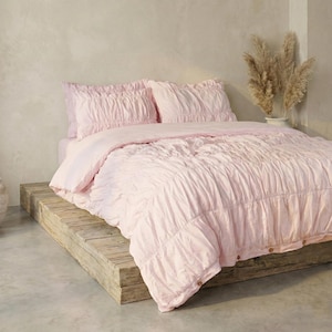 May include: A bed dressed in light pink bedding, featuring a comforter and two pillowcases. The bedding has a textured, gathered design. The bed is on a rustic wooden platform. Dried ornamental grass in a white vase is in the background.