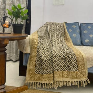 May include: A beige and black patterned throw blanket with a geometric design and a tan border with fringe. The blanket is draped over a sofa with blue cushions.