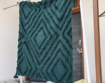May include: A dark teal throw blanket with a geometric diamond pattern. The blanket is hanging on a metal rod, showcasing its textured design and tassel accents. The blanket's dimensions are not visible.