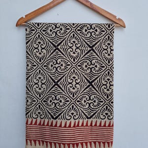 May include: A beige and black patterned textile hanging on a wooden hanger. The fabric features a repeating geometric design with floral elements. The bottom edge has a red and beige striped border. The textile is likely a scarf or shawl.