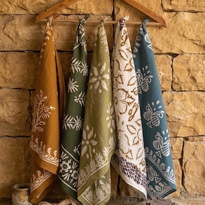 Peut inclure: Cinq torchons de cuisine à motifs dans les tons moutarde, vert olive et sarcelle sont suspendus à un cintre en bois. Chaque torchon présente un motif floral ou géométrique blanc unique. Un petit pot en céramique et de la ficelle sont visibles.