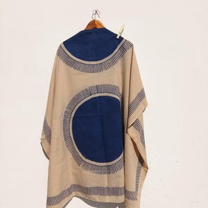 Op de afbeelding: Een beige en marineblauw patroon kimono hangt aan een houten kleerhanger tegen een witte muur. De kimono heeft grote marineblauwe cirkels met stralende lijnen en een effen marineblauwe pas. Het kledingstuk is vastgezet met een wasknijper.
