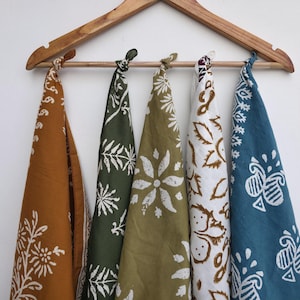 May include: Five patterned bandanas in shades of brown, green, and blue hang from a wooden hanger. Each bandana features a unique white floral design. The bandanas are knotted at the top.