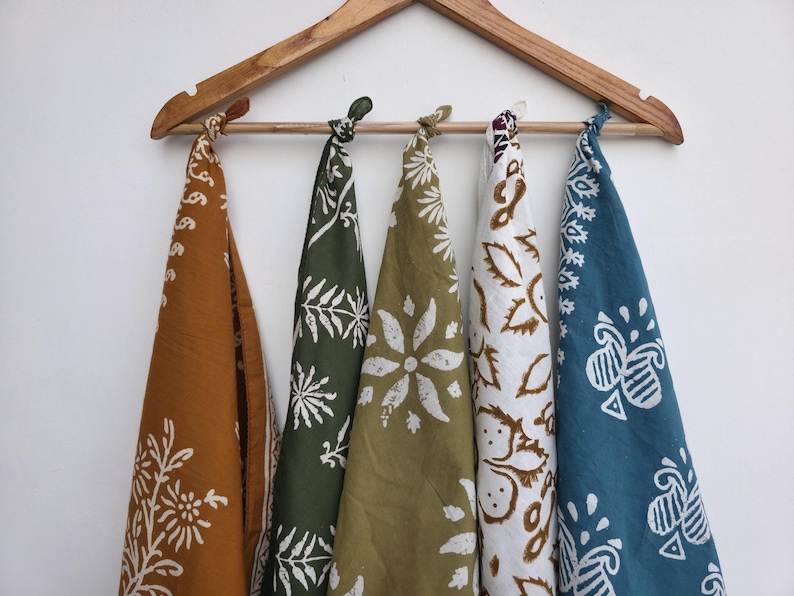 May include: Five patterned bandanas in shades of brown, green, and blue hang from a wooden hanger. Each bandana features a unique white floral design. The bandanas are knotted at the top.