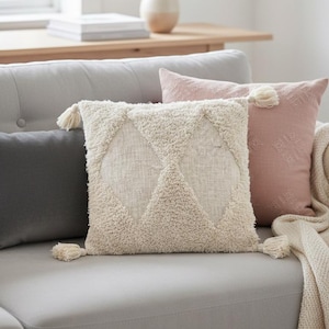 Könnte beinhalten: Ein graues Sofa mit mehreren dekorativen Kissen. Die Kissen umfassen ein strukturiertes cremefarbenes Kissen mit einem Rautenmuster, ein staubrosa Kissen und ein dunkelgraues Kissen. Eine cremefarbene Decke ist über das Sofa drapiert.